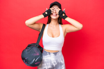 Young beautiful sport woman with sport bag isolated on red background covering eyes by hands
