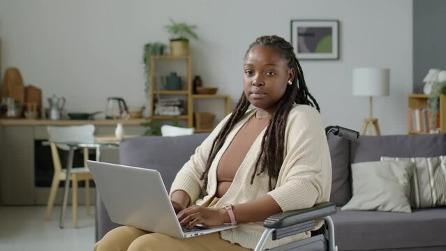 Portrait of young African American woman with disability sitting in wheelchair, holding laptop and posing for camera in living room at home