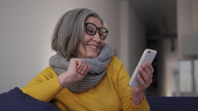 Extremely happy excited woman listening to favourite music on smartphone application and dancing, sitting on sofa, having fun enjoying performance online, excitement