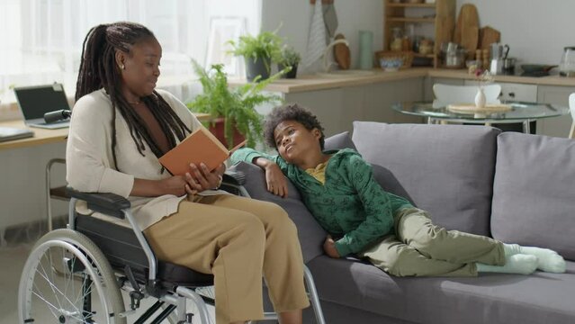 African American mother in wheelchair reading book to little son chilling on sofa while spending time together at home