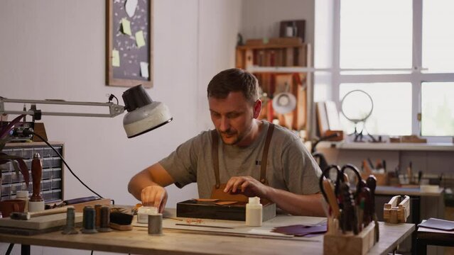 Leathersmith adjusts table lamp for comfortable leather processing in workshop. Bearded master crafts item in small studio. Workplace of handcraft designer