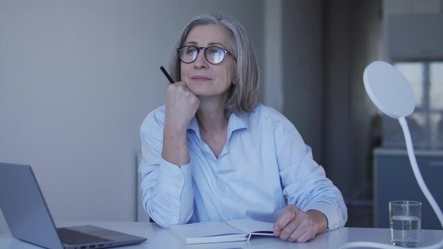 Attractive senior woman writer taking notes to new chapter of her book, dreaming and inspiring herself, writing down her thought to notebook, smiling while imaging happy end