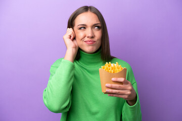 Young caucasian woman holding fried chips on purple background frustrated and covering ears