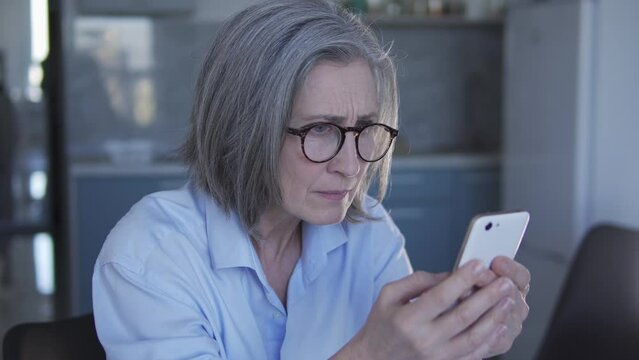 Gray-haired woman freelancer reading on smartphone feedbacks about her work, shocked with bad comments, upset because of dishonest clients, got into a scam