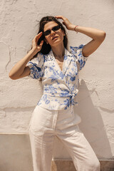 Fashionable young caucasian woman posing near light wall in sunny weather outdoor. Model with brunette wavy hair wears casual clothes and sunglasses. Concept enjoying sunshine.