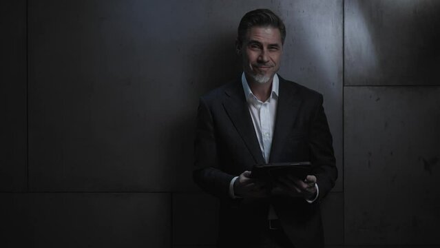 Portrait of happy mature businessman with tablet computer. Entrepreneur standing in front of dark  wall. Confident older, middle aged, mid adult, man in his 40s or 50s in business casual.