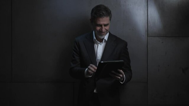 Portrait of happy mature businessman with tablet computer. Entrepreneur standing in front of dark  wall. Confident older, middle aged, mid adult, man in his 40s or 50s in business casual.