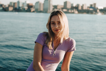 Portrait photo of a woman by the water overlooking the city. The blonde woman smiles and looks at the camera. Summer time. Sunny day. Golden time
