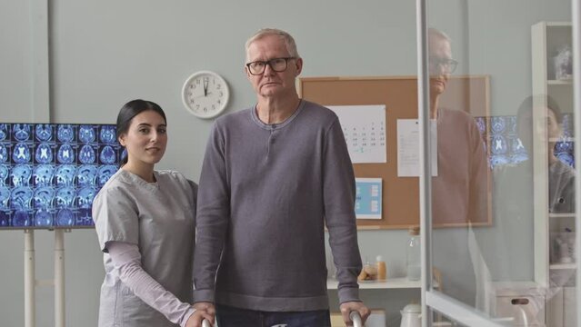 Medium portrait of young female nurse and senior man with disability leaning on walkers looking at camera standing at modern doctors office