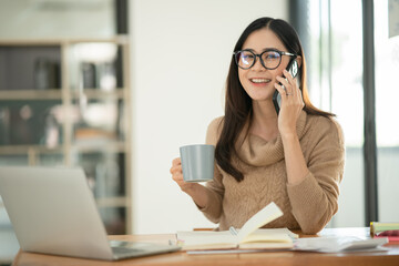 Obraz premium Asian businesswoman holding coffee mug, documents standing and talking on phone with customer.