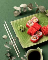 Sushi rolls on green plate on green cloth background with a green branch, with bamboo sticks and soy sauce. Vegetarian dish. Healthy eating. Japanese cuisine. Top view.