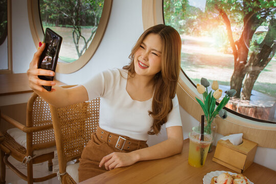 Portrait of cool cheerful girl having video call with lover holding smart phone in hand shooting selfie on front camera. Good mood lady with expansive smile enjoying started weekends in the cafe.