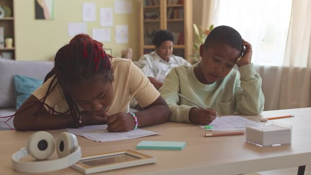 Two African American 2nd grade siblings doing Math worksheets at desk in living room while staying at home with their grandma sitting on couch in background