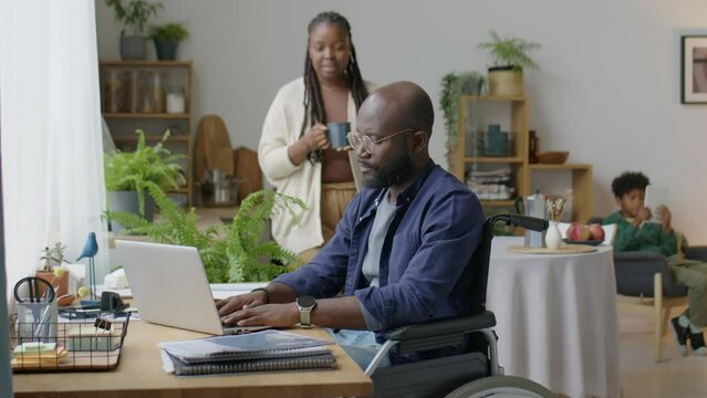 Black Man In Wheelchair Working Remotely On Laptop From Home While Wife Bringing Him Cup Of Tea