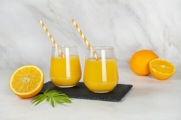 Freshly squeezed orange juice in two glasses with tubes on a stylish black slate tray. Orange slices and a green tropical plant on a light marble background. Healthy food and lifestyle, fruit bar