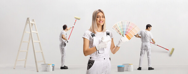Female house painter holding a color swatch palette and pointing and other workers painting