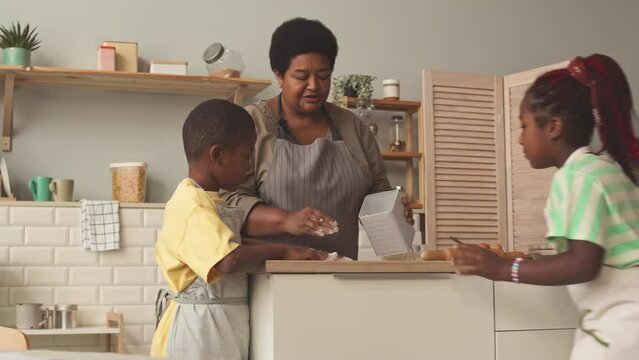 African American grandmother strewing some flour on kitchen table while making dough with grandchildren at weekend