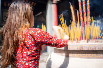 Vietnamese girl in traditional long dress or Ao Dai dress is praying with incense stick in the...