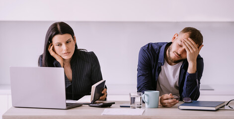 Overworked young businesswoman in suit works using laptop holds diary sitting at desk with sleepy young man with closed eyes touching forehead. Fatigue, exhausted. Couple in troubles. Debts. Deadline.