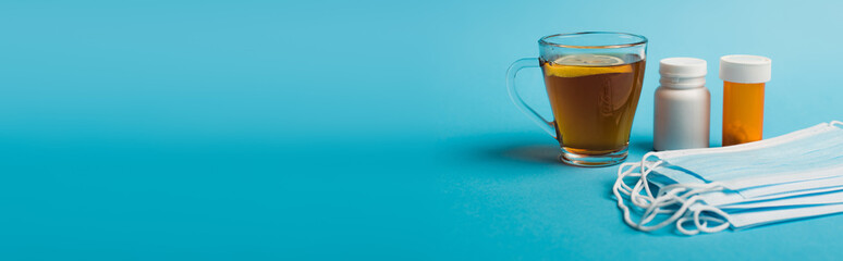 Medical masks near pills and cup of tea with lemon on blue background, banner.