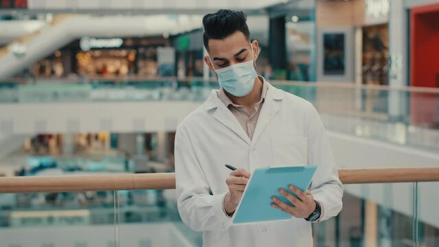 Ethnic Indian inspector scientist doctor medical male worker man writing in papers documents expertise check health safety rules supervisor with medical mask robe inspecting shopping mall write data