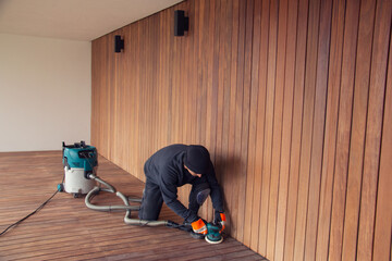 Professional deck worker kneeling while sanding wooden deck with orbital power sander collecting...
