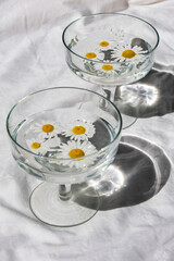 Glass glasses with water where daisies are floating cast a shadow on the white tablecloth. Vertical image.