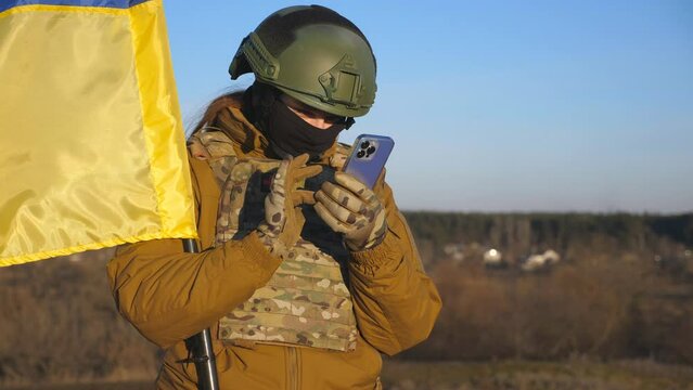 Female Ukrainian Army Soldier Using Smartphone To Read News Or Messaging With Family. Girl In Military Uniform And Helmet Holding Waving Flag Of Ukraine. Victory Against Russian Aggression. Close Up