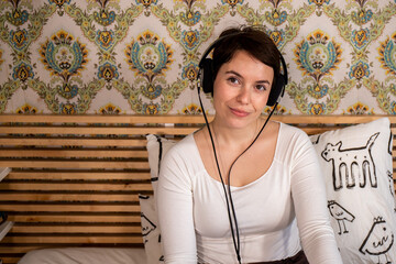 Beautiful woman listens to music in headphones on the bed. The woman looks towards the camera while listening to her favorite song of hers about her.
