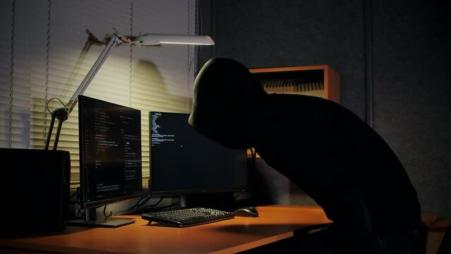A hooded computer hacker turns on a desk lamp and sits down at his workplace, then types in a code and launches a program to hack websites and infect government resources with computer viruses.