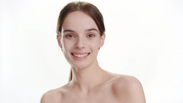 Body care concept | Close-up shot of gorgeous young slim Caucasian woman with long brown hair strokes her shoulder turning her head to the camera and smiles widely for it on white background