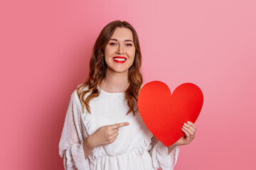 Valentine's Day concept. Caucasian smiling girl holding valentine card pointing finger at big paper heart isolated on pink background.