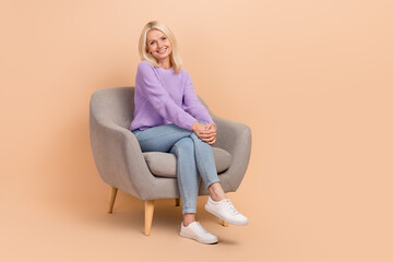 Fototapeta na wymiar Full size photo of cheerful positive woman dressed purple sweater jeans sit on armchair hands on knees isolated on beige color background