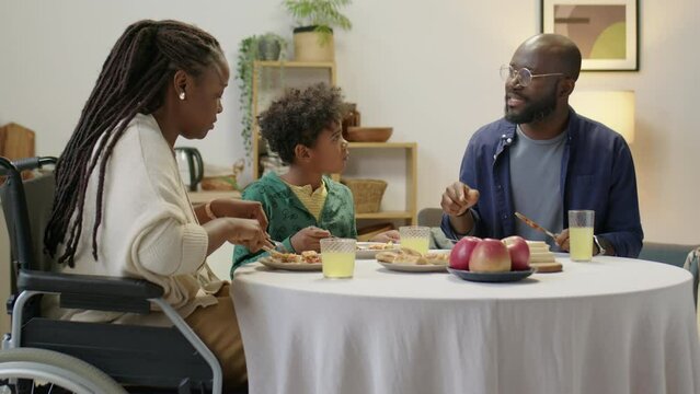 Medium shot of African American woman in wheelchair eating dinner with little son and husband in evening at home