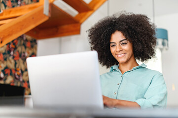 Fototapeta na wymiar Concentrated young multiracial businesswoman in casual wear sitting at the desk in the home office, working remotely on the laptop. Smiling freelancer woman typing, answering emails
