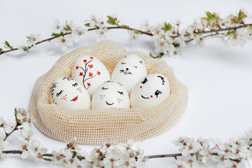 Fototapeta na wymiar Defocused focus on foreground sprigs of flowers. Easter beautifully painted eggs in eco-friendly mesh bag on white table. Funny spades, flowers, cat's face are painted on eggs. Copy space
