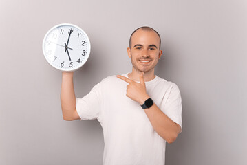 Balding smiling man is pointing at the white round clock he is holding over grey background.