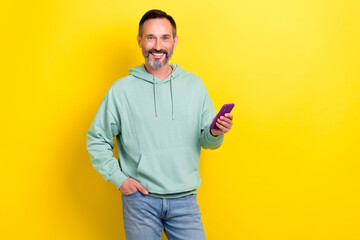 Photo of satisfied technology gadget user middle age man wear khaki hoodie hold phone browsing new dating app isolated on yellow color background