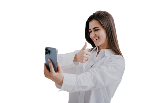 Beautiful  brunette girl in white shirt, makes selfie over transparent background. Successful blogger  welcoming subscribers. Cheerful student holds phone makes video call shows thumb up gesture