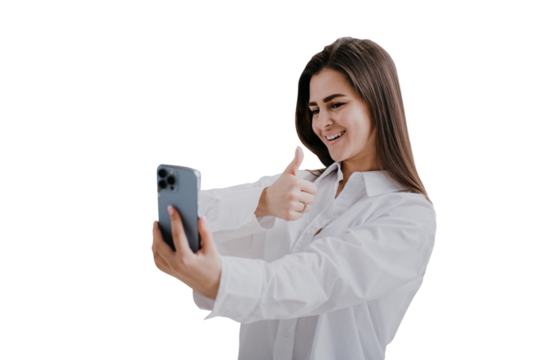 Beautiful brunette girl in white shirt, makes selfie over transparent background. Successful blogger welcoming subscribers. Cheerful student holds phone makes video call shows thumb up gesture