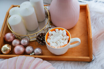 Mug of coffee with marshmallows on a wooden tray, white candles. Christmas decor element in pink color.