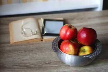 Open book, e-reader, reading glasses and bowl of apples on the table. Bookshelf in the background. Selective focus.