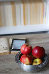 Open book, e-reader, reading glasses and bowl of apples on the table. Bookshelf in the background. Selective focus.