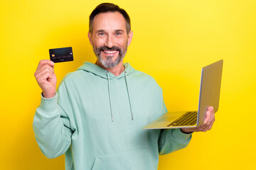 Portrait of toothy beaming positive man dressed green hoodie hold laptop demonstrate debit cart isolated on yellow color background