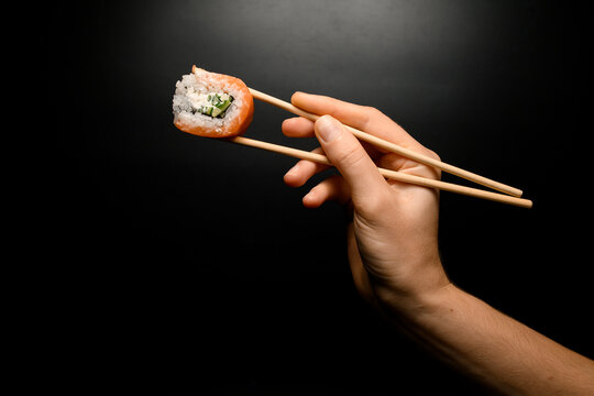 female hand accurate holding Philadelphia roll with bamboo chopsticks on dark background.