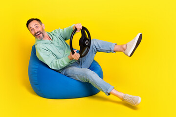 Full length photo of funny excited man wear green sweatshirt driving auto having fun sitting beanbag isolated yellow color background