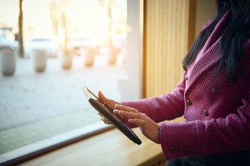Cropped view woman in stylish casual wear, using digital tablet, works remotely on new action for her business project, plans meetings with investors and partners, standing by windowsill in her office