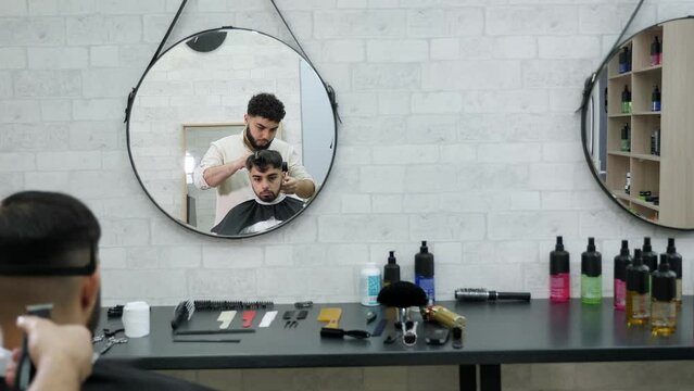 A young handsome man cuts a man's hair in a modern barbershop, the client is sitting in a black leather chair in front of a mirror.