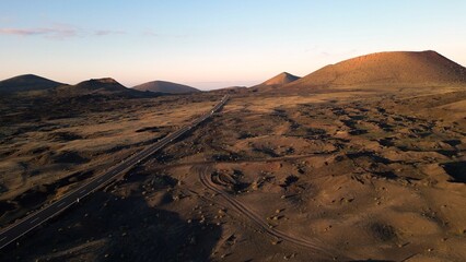 Fototapeta na wymiar Europe, Spain, Lanzarote, Canary Islands - Drone aerial view of amazing volcanic landscape at sunset in Tymanfaya national park with road passing between black lava 