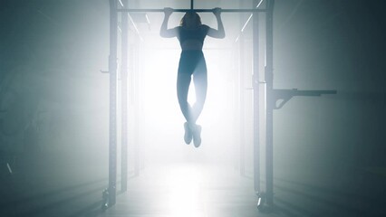 Silhouette of a sporty woman lifting her body while exercising at the gym studio. Female athlete hanging from a bar by her hands and pulling herself upward. High quality 4k footage